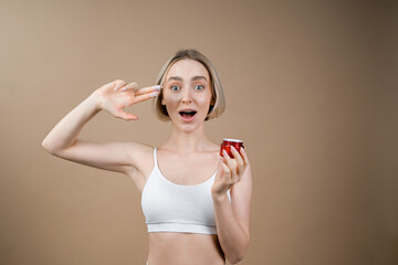 Young woman with healthy face complexion holding small red container with skincare cream. Model in white sport top posing on beige background, advertising cosmetic products.