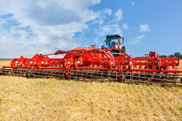 Universal red harrow as a hitch on an agricultural tractor against the background of a compressed yellow wheat field.