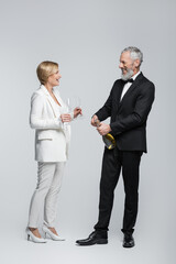 Positive mature groom opening champagne near bride with glasses on grey background.