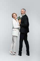 Cheerful mature bride holding wedding bouquet near groom looking at camera on grey background.