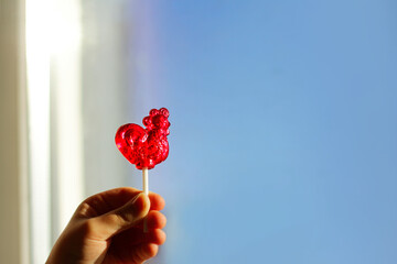 Transparent lollipop red cockerel on a stick. Bright blue background sky.