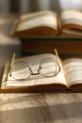 Open book and reading glasses on the table, illuminated by sunlight. Stack of vintage books in the background. Selective focus.