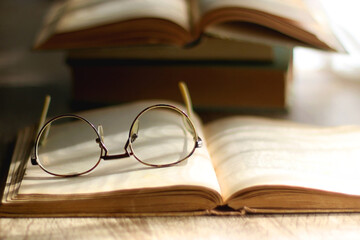 Open book and reading glasses on the table, illuminated by sunlight. Stack of vintage books in the background. Selective focus.