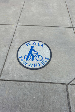 Round sign with a picture of a person near a bike painted on a cement sidewalk that says, walk your wheels