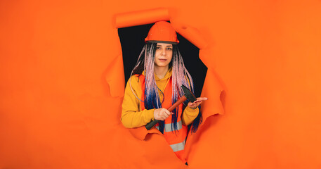 Female construction worker with hammer sticking out of hole of orange background. Young woman in overalls slaps tool on hand and looking at camera.