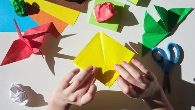 Children's hands do origami from colored paper on white background. Origami paper. DIY. Various school supplies. Concept of children's creativity, back to school.	
