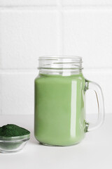 fresh green smoothie in glass bottle and spirulina powder with spoon on ceramic tile background. self care and healthy lifestyle. copy space
