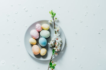 Top view Colored Easter eggs and blooming tree branch on the gray plate with flower petals on the white table. Happy Easter and springtime card. Minimalism and simplicity. Selective focus. Copy space.