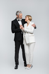 Full length of grey haired groom hugging bride in suit on grey background.