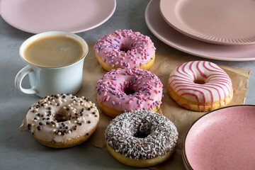 Several donuts lie on craft parchment paper on gray surface. Porcelain cup of coffee and pink plates stands next to donuts laid out on table. Selective focus.