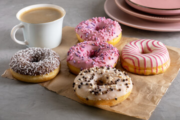 Several donuts lie on craft parchment paper on gray surface. Porcelain cup of coffee and pink...