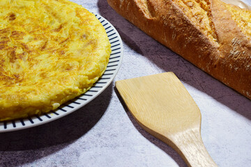 Potato omelette, typical of Spain, on a porcelain plate, stone background, along with a wooden utensil and bread.