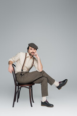 full length of man with mustache in vintage style clothing and hat sitting on wooden chair on grey.