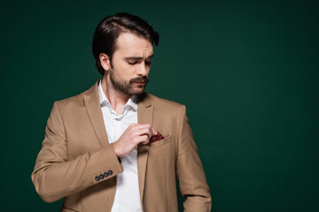 young man with mustache adjusting handkerchief in pocket of blazer on dark green.