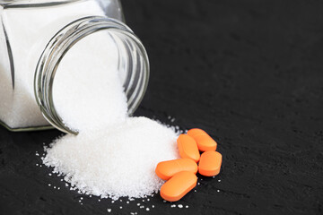 Glass jar with sugar and orange pills on a black background.Diabetes. Scattered white sugar on a black table. Copy space. Place for text.Extra calories
