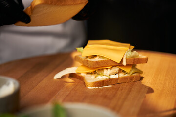 Close up view of vegetarian sandwich with salad, cheese and egg on cutting board. Crop view of hand putting toast on top of sandvich in restaurant kitchen. Concept of food. 