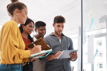 Churning out great ideas with the smartest tools. Shot of a group of businesspeople using a digital tablet while brainstorming on a glass wall in an office.