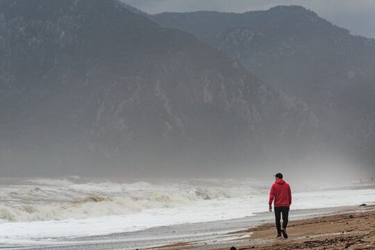 A man walks along the beach on the Mediterranean Sea in Turkey, Antalya, against the backdrop of big waves and mountains, cloudy weather, rain, huge waves.