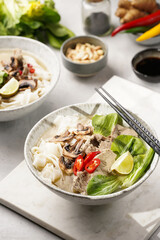 Pho Bo vietnamese soup with beef, rice noodles, pak choi and mushrooms in a grey bowl on marble board, peanuts, soy sauce, chilli