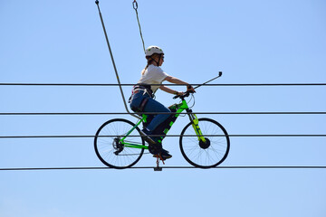 A young woman rides a ride on a bicycle on ropes above the ground. Against the background of the sky