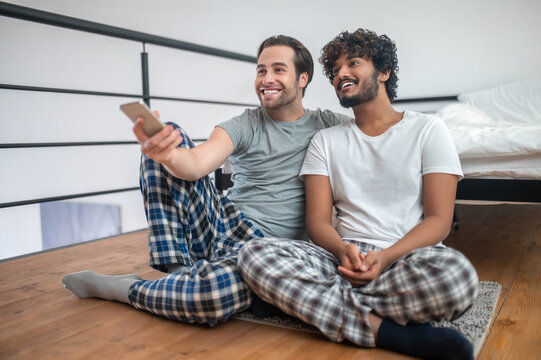 Joyful gay couple sitting on the floor mat during channel-surfing - Powered by Adobe