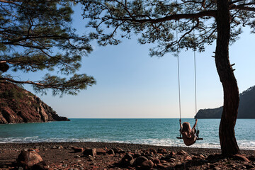 A girl swings on a swing on the shores of the Mediterranean Sea, on a clear sunny day, against the backdrop of mountains, blue water, rest, Antalya, Turkey