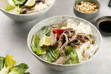 Pho Bo vietnamese soup with beef, rice noodles, pak choi and mushrooms in a grey bowl, peanuts, soy sauce, chilli