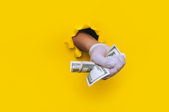 A doctor hand holds money (dollars) in white medical glove. Yellow torn hole paper background.The concept of treatment fees, bribes, illegal surgery. Copy space.