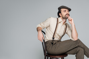 pensive man with mustache in retro style clothing and hat sitting on wooden chair isolated on grey.