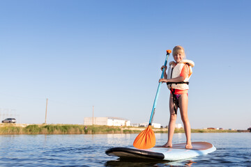 Cute little caucasian blond child girl enjoy having fun stand on sup board surfing at freshwater...