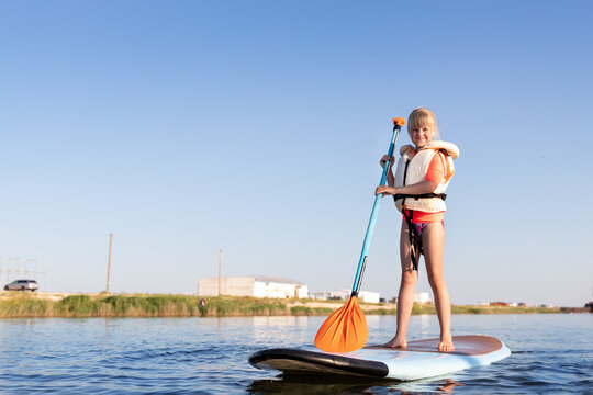 Cute little caucasian blond child girl enjoy having fun stand on sup board surfing at freshwater pond lake or river hot sunny day. Summer outdoors child healthy sport recreation activities