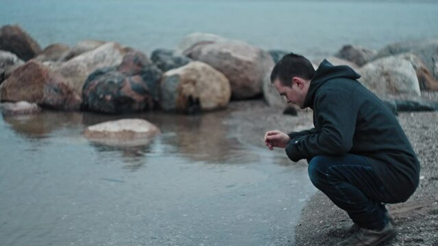 A young guy comes to the seashore and touches the water, checking the temperature of the water he is dressed in casual dark clothes the action takes place on the ocean on a gloomy autumn day copyspace