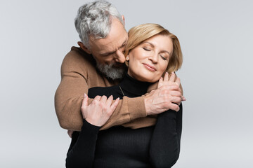 Grey haired man hugging wife with closed eyes isolated on grey.