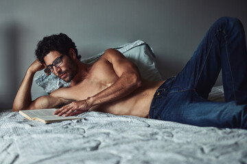 He enjoys reading novels over the weekend. Cropped shot of a handsome young shirtless man reading a book on his bed at home. - Powered by Adobe