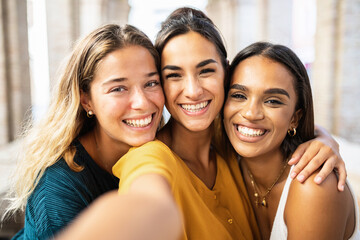 Three multi ethnic young female friends laughing together taking selfie outdoors - Multiracial...