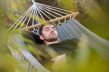 Jeune homme de 20 ans qui fait la sieste dans son jardin, allongé dans un hamac. Au premier plan, il y a des plantes.