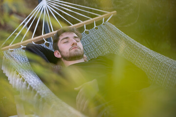 Jeune homme de 20 ans qui fait la sieste dans son jardin, allongé dans un hamac. Au premier plan, il y a des plantes. - obrazy, fototapety, plakaty