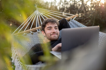 jeune homme, étudiant ou employé, homme d'affaire travaillant à distance avec son ordinateur portable, allongé dans un hamac, dans son jardin