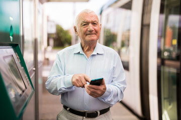 Senior European man standing on tram stop and using his phone.