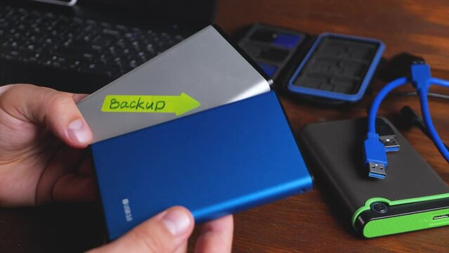 External hard drive in man's hand on desk background. Black laptop, USB cable. Data backup in information technology. Copy of computer data. Restore original after data loss event