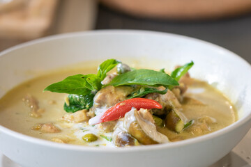 Close up to Green Curry with Chicken garnished on top with pepper and basil was served with white bowl put on wicker cream color placemat.