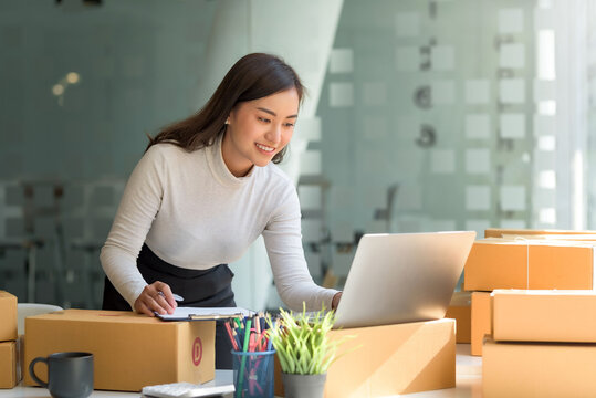 Happy young Asian business owner woman prepare parcel box and check online orders of product for deliver to customer on laptop computer. Shopping Online concept.