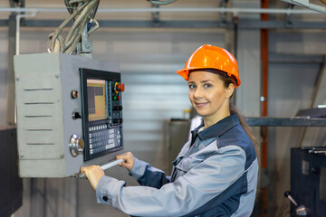 a woman in overalls and a helmet works on a machine tool at a factory at the control panel. the specialist looks at us, smiles. factory, production. portrait