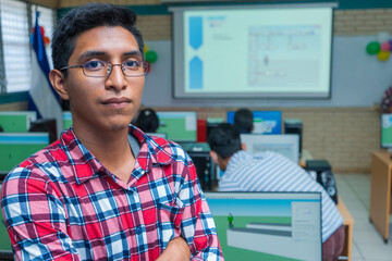 Portrait of a young Latin Central American man in glasses with his arms crossed and a slight smile standing in a classroom full of computers - obrazy, fototapety, plakaty