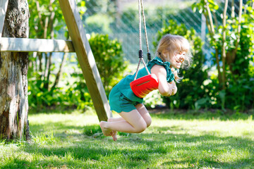 Happy little preschool girl having fun on swing in domestic garden. Healthy toddler child swinging on sunny summer day. Children activity outdoor, active smiling kid laughing - obrazy, fototapety, plakaty