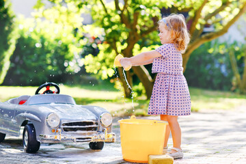 Cute gorgeous toddler girl washing big old toy car in summer garden, outdoors. Happy healthy little...