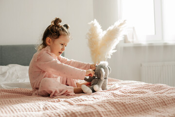A cheerful little girl on the bed after waking up plays with her toy elephant