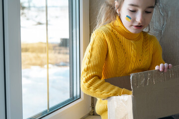 A child on the window with a humanitarian aid box with the flag of Ukraine painted on his cheek. A...