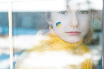 A sad child look at the window with the flag of Ukraine painted on the cheek, worries and fear. Humanitarian aid to children, world peace, security.