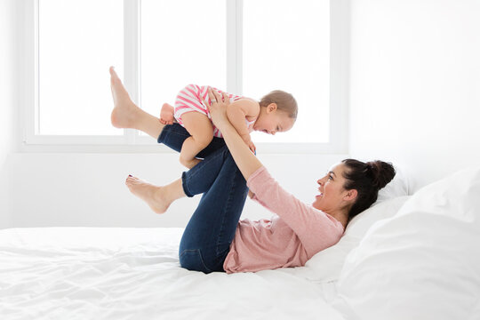 Mother lying on white bed making her baby girl fly on her legs